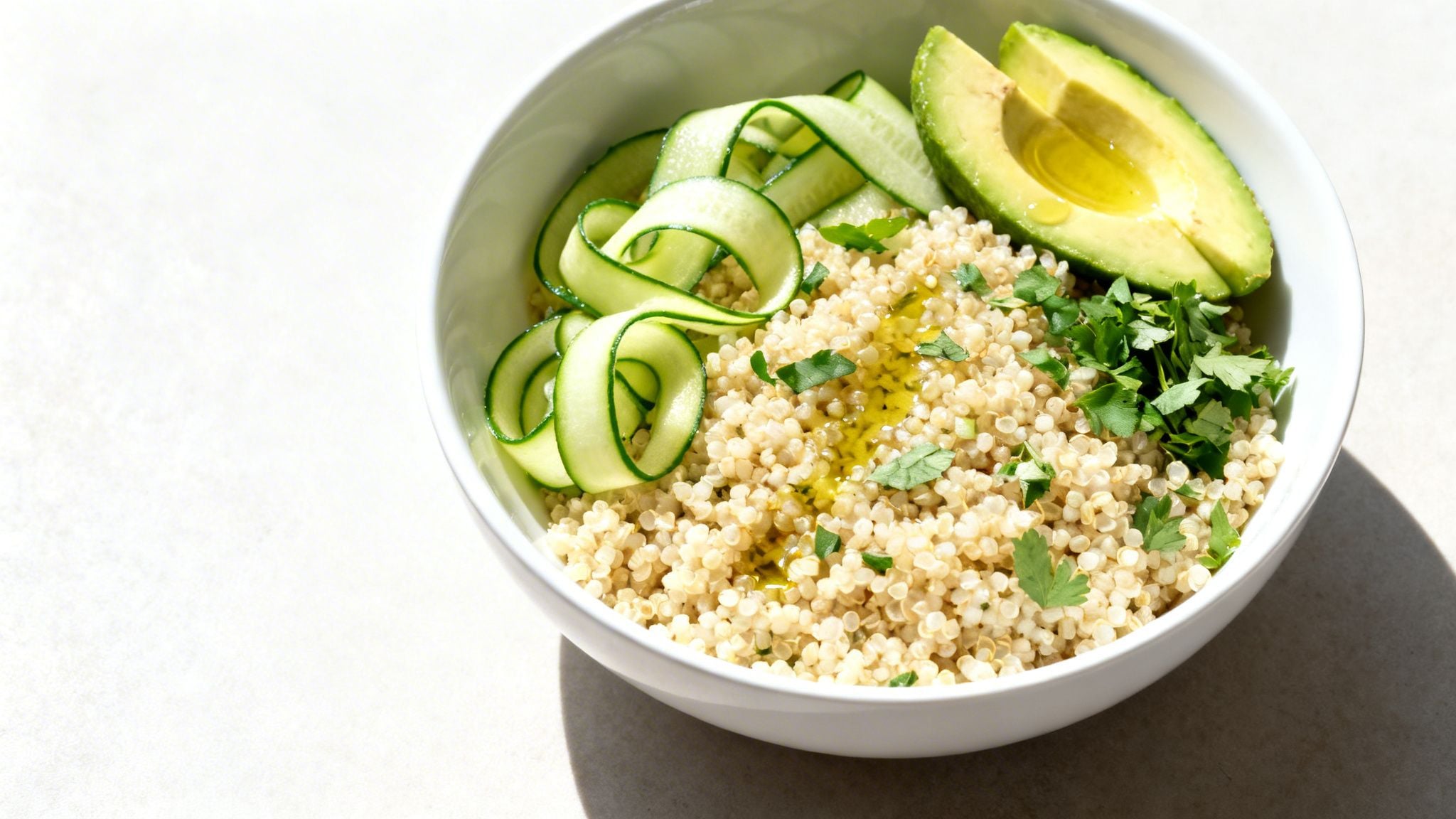 A healthy quinoa bowl topped with sliced avocado, ribbons of fresh cucumber, and chopped herbs with olive oil.