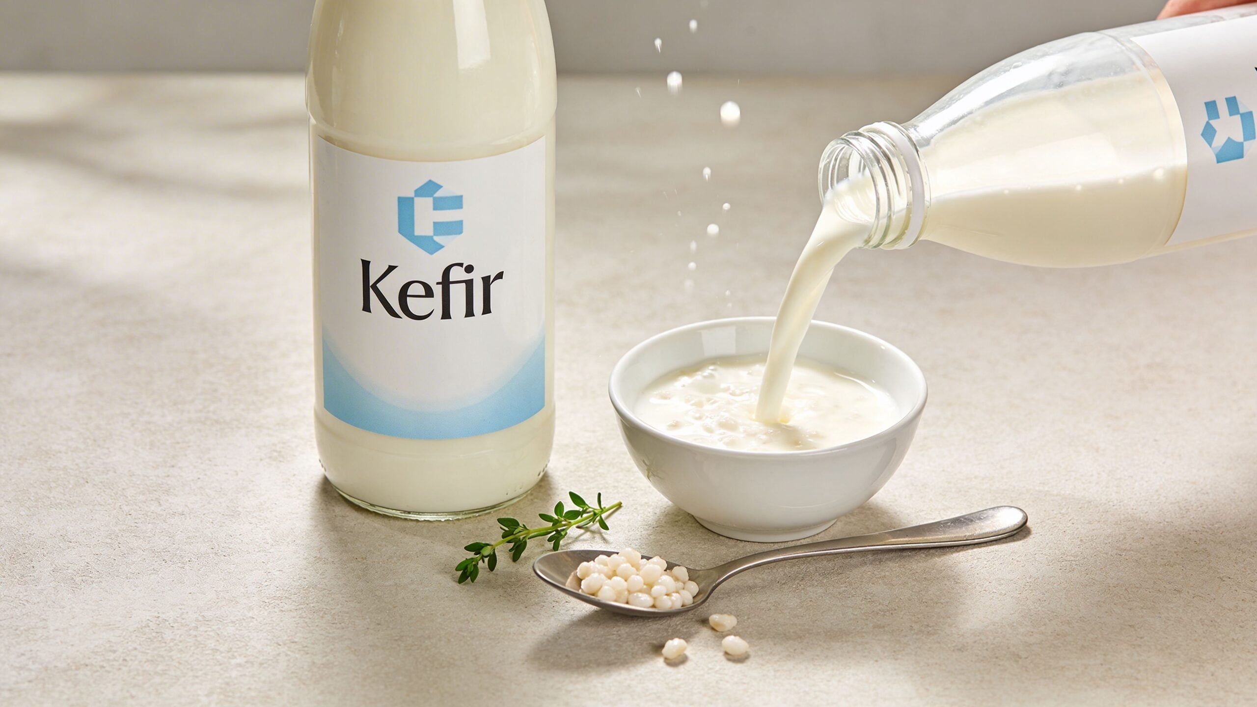 A bottle and bowl of kefir being poured, with kefir grains on a spoon and fresh thyme herbs.