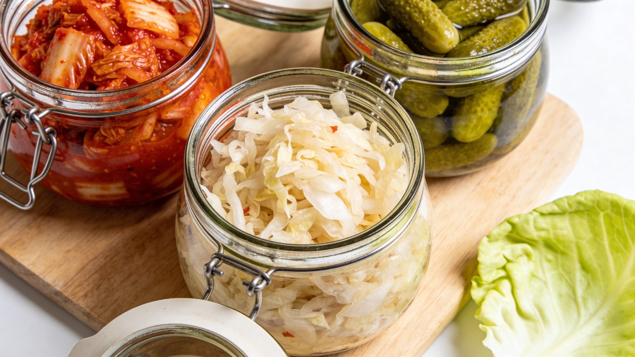 Three glass jars filled with fermented foods like kimchi, sauerkraut, and pickles on a wooden board.