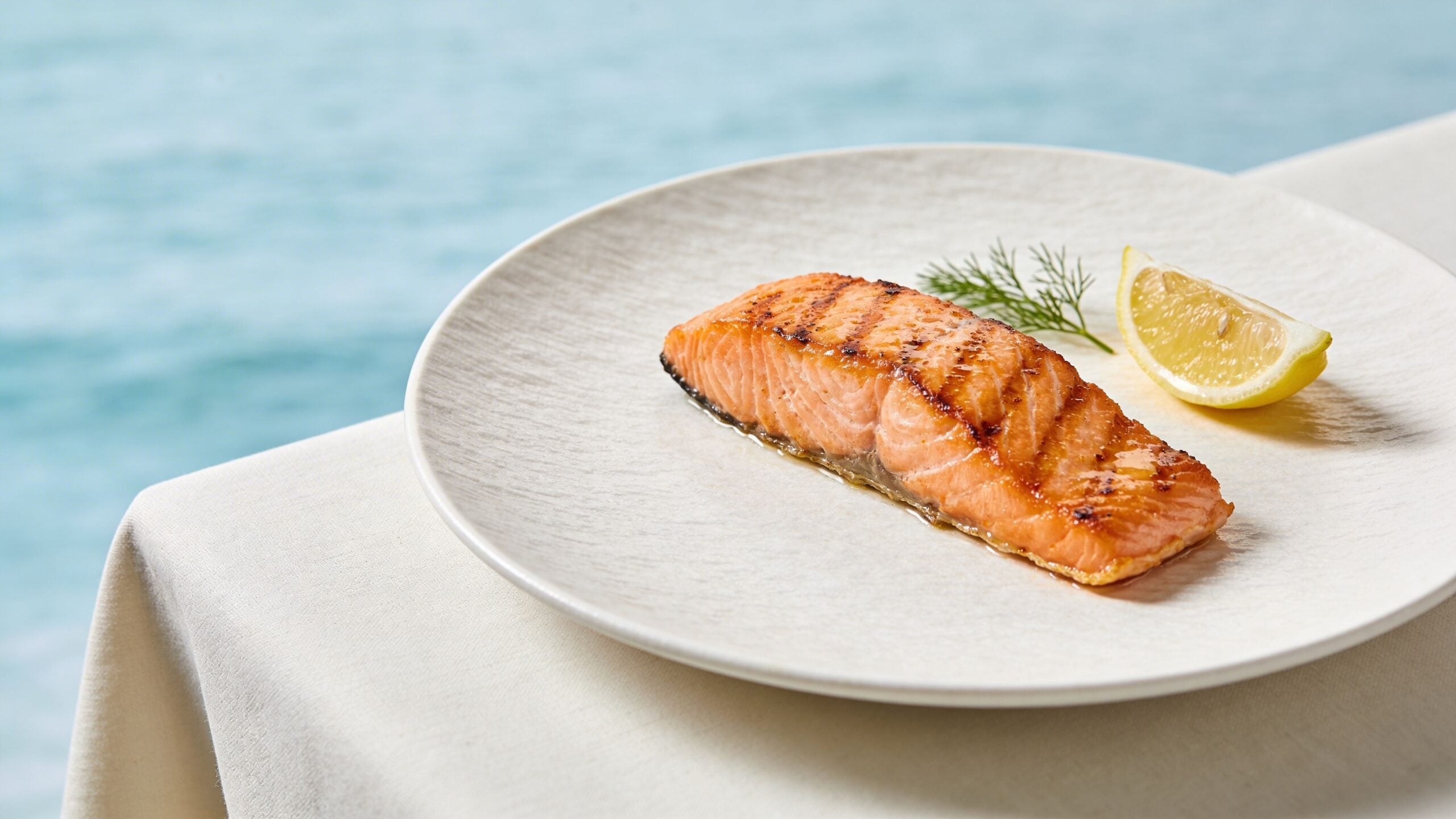 A grilled salmon fillet served on a white plate with fresh dill and a slice of lemon.